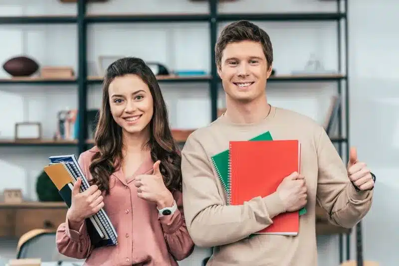 Két mosolygó diák füzetekkel hüvelykujjat mutat a Személyes Angol Tanfolyam Budapesten óráján.